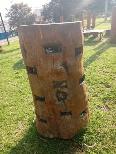 A thick, carved wooden stump stands in a grassy field. Square and rectangular holes punctuate its surface, some filled with dark, possibly resinous material. In the background, more wooden structures rise from the grass, including upright posts and a picnic table. The sunlight casts shadows on the uneven green lawn. A building and trees are visible in the distance.