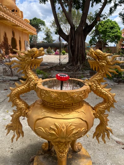 In the city of Đăk Mar, Vietnam, a large, ornate golden incense burner stands in the foreground of an outdoor scene. The burner is fashioned with two elaborate golden dragon statues acting as handles, their heads facing outward from the central basin. The basin contains numerous thin, dark, vertically oriented incense sticks embedded in a substrate, with a red, lotus-shaped solar-powered light fixture positioned centrally among them. The burner's body features sculpted lotus petals and rests on three legs, each terminating in a dragon head motif. The immediate ground is concrete. To the left, a multi-tiered ochre-colored temple building with intricate architectural details and several white statues is partially visible. In the midground, a large, gnarled tree with exposed roots dominates the center-right, beyond which lies an open area with trimmed bushes, some displaying pink flowers, and additional white statues. The background features a partially cloudy sky and dense foliage. No individuals are present in the scene.
