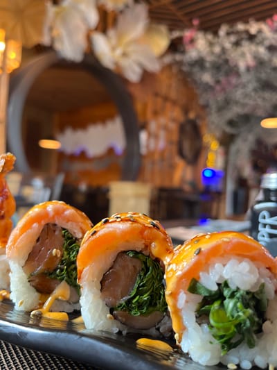 The image presents a close-up, shallow-depth-of-field view of three sushi rolls arranged on a dark, rectangular serving plate, positioned on a dark, woven placemat. Each roll is composed of white rice, filled with green leafy vegetables and a piece of brown fish, topped with a slice of raw salmon, drizzled with an orange-yellow sauce, and sprinkled with white and black sesame seeds. Additional sauce is visible on the plate.

The background, significantly blurred, depicts a restaurant interior. A large, dark, arched entryway occupies the mid-left, revealing a warm-toned room with a distinct, jagged white decorative element against a darker wall. To the right of the arch, a wall features vertical wooden slat paneling. Blurred white and pink floral decorations are suspended throughout the ceiling area. Multiple diffused yellow light sources and a prominent blue light are visible as bokeh effects. On the far right, a dark bottle with partial white lettering, "PPER," is present on a surface.

This scene is located within the Tokyo Grill & Sushi restaurant in Bad Sobernheim, Germany.