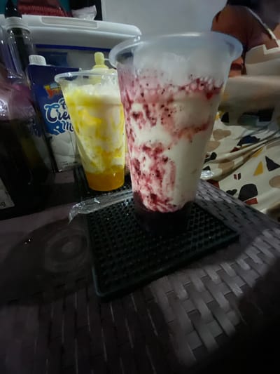 Two plastic cups containing layered beverages are displayed on a dark, textured surface, likely a table or tray. The cup on the right is filled with a creamy white drink swirled with red streaks, possibly a fruit puree or syrup. The cup on the left contains a yellow beverage layered with white, suggesting a mixed drink like a mango smoothie or shake. In the background, a blue carton with the word "Cream" is visible, along with a dark bottle. The setting appears to be indoors, possibly a home or a food establishment, and the lighting is dim, creating shadows. The overall impression is of a prepared dessert or beverage, ready for consumption.