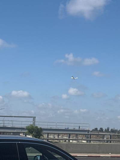 A small white airplane is flying in a clear blue sky with scattered white clouds. In the foreground, a dark-colored car is partially visible, suggesting the photo was taken from within a vehicle. Behind the car, a multi-level overpass or bridge structure is visible with railings and supports. Trees and other vegetation are present in the background, behind the bridge structure, and what appears to be a road or ground level area. The scene is consistent with an outdoor environment in an urban or suburban setting, possibly along a roadway. The presence of the airplane suggests it is in an area with air traffic, which could be near an airport. Given the prompt's mention of Kfar Shmaryahu, Israel, this scene is situated within that locality.