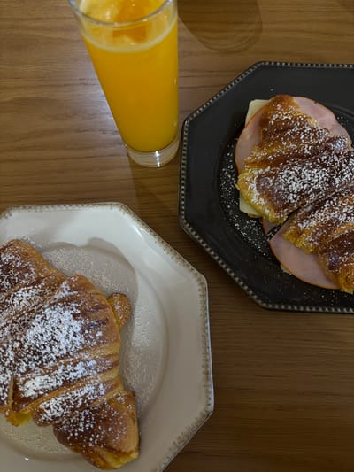 An overhead view captures a breakfast or snack arrangement set on a light brown wooden table in Viseu, Portugal. A tall, clear glass, containing fresh orange juice with visible pulp, is positioned towards the upper-left, casting a shadow to its right. To the upper-right, a dark grey octagonal plate with a raised, beaded rim holds a croissant sliced open and filled with two slices of pink ham and a light yellow cheese slice. This savory croissant is dusted with white powdered sugar. To the lower-left, a light cream-colored octagonal plate, also with a raised, beaded rim, presents a plain croissant, similarly dusted with white powdered sugar. No individuals are visible in the scene.