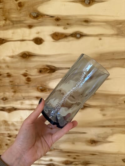 A human hand holds a transparent, smoky-tinted glass against a light wooden background. The wood features prominent vertical grain patterns and numerous dark knots, suggesting a plywood panel or wall. The hand, with dark, glittery nail polish visible on the index finger and a plain gold-colored ring on the ring finger, holds the glass tilted. Embossed or debossed text is visible on the side of the glass. Due to the glass's transparency and angle, the Cyrillic text is mirrored and slightly distorted, but it clearly reads "СОБСТВЕННОЙ ИСТОРИИ" (Sobstvennoy Istorii), which translates to "OWN HISTORY" or "PERSONAL HISTORY". The scene appears to be indoors. The location context is Kuda, Russia.