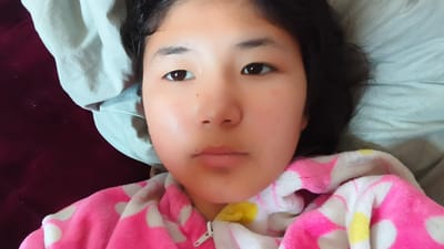 This is a close-up, low-angle selfie of a young East Asian girl lying down in bed. She has dark hair, dark eyes, and fair skin with a reddish hue on her cheeks. She is wearing a pink and white polka-dot hooded robe with a zipper. The background shows a light blue blanket and a dark red surface, possibly a bedspread or furniture. The lighting suggests it could be daytime, with natural light illuminating her face. The girl's expression is neutral, and she appears relaxed, as if waking up or getting ready to sleep. There is no visible text in the image. The setting is likely a bedroom in Uchkun, Kyrgyzstan.