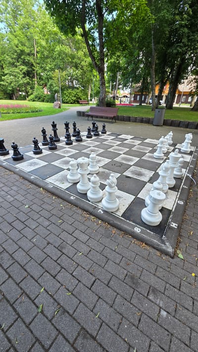 The image captures a life-size chess board set up on a paved area in a park, likely in Palanga, Lithuania. The chess board is positioned in the foreground, with black and white oversized chess pieces arranged on it. The white pieces are on the right side of the board, while the black pieces are on the left. The chess board and pieces are weathered, indicating they are outdoor furniture. The park setting includes lush green trees, a well-maintained lawn, and walking paths made of grey pavers. In the background, there are benches, a trash receptacle, and buildings that suggest a public space within a town or city. The overall impression is of an outdoor recreational area designed for leisure activities.
