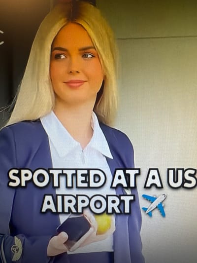 The image shows a young woman with blonde hair and light skin, wearing a blue uniform with a white collared shirt. She has makeup on, including eyeliner and lipstick. Her hair is styled in loose waves. She is lying down and looking towards the camera with a slight smile.

There is text overlaid on the image that reads "SPOTTED AT A US AIRPORT". There is also a small blue and white airplane graphic next to the text. A part of a dark surface is visible in the foreground, and the background appears to be a lightly textured wall.

The image has a slightly grainy appearance, suggesting it may be a screenshot or a photo taken with a flash. The orientation of the image is such that the woman's head is on the left and the text is on the right, running vertically.

The woman's attire and the text suggest she might be an airline employee or someone involved with air travel. The emotions conveyed are relaxed and friendly. The specific location context of Chorzów, Poland, does not appear to be directly reflected in the visual elements of the image itself.