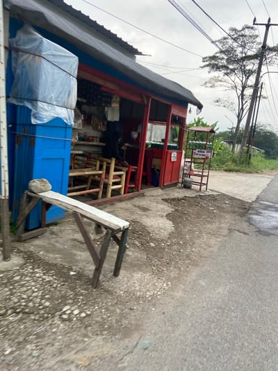 The image shows a small shop with a red painted exterior in Kecamatan Margaasih, Indonesia, located at coordinates (latitude: -6.963, longitude: 107.538). There is one person, partially obscured, seated in the shop. The shop has a blue painted door and is open to the front. There are several wooden chairs and tables in the shop. A wooden bench is sitting outside the shop. The shop is situated on a gravel path leading to a paved road with a tree line on the right side. The shop has a small metal shelf displaying goods for sale. The shelf has a sign with the words "Bensin Pertalite" written on it. The area is partially obscured by a power line, which is supported by a wooden pole. There are numerous power lines visible in the sky. The sky is a light gray and overcast. The road appears to be in need of repairs, with many potholes visible.