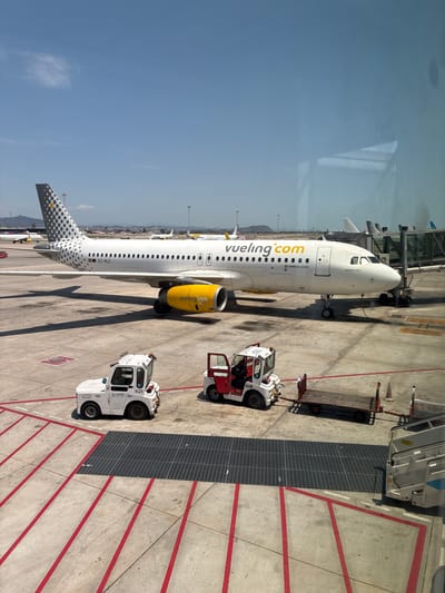 The image shows an airplane at an airport gate on a sunny day. The airplane is white with a grey and yellow pattern on its tail and yellow engines. The airline name "vueling.com" is visible on the side of the plane. In front of the plane are two small airport vehicles, a white tug and a red and white tug, and a baggage cart. The airport tarmac is marked with red and white lines. In the background, other airplanes and airport structures can be seen. The scene is observed through a window, with reflections visible on the glass. The location is not Großbeeren, Germany.