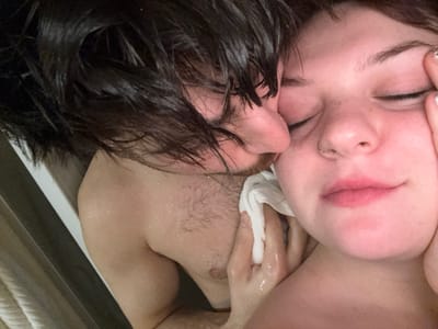 A close-up, intimate shot shows two people in the shower. The person on the left has dark, wet hair and is turned toward the other person, who has their eyes closed. A white cloth is held by a hand, possibly drying their face. Water droplets glisten on their skin, indicating they are wet. Their faces are close together, creating a feeling of closeness and affection. The scene appears to be a tender moment shared between the two individuals.