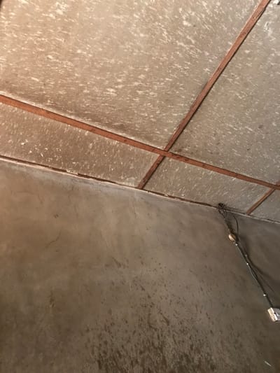 The image displays an interior corner of a room, likely a utilitarian space such as a storage area or a basement, situated in Oba-Ile, Nigeria. No people are present in the scene.
The surface on the left side of the frame consists of light grey or beige panels, possibly made of concrete or a similar material, which are separated by dark reddish-brown strips arranged in an irregular grid pattern. These panels feature a prominent mottled texture with white speckled areas, suggesting either an uneven paint job, dried water stains, or a specific textured finish.
The surface on the right is a plain, untextured wall of light grey, characteristic of concrete or plaster. Along the upper section of this wall, near the corner, black electrical wires are visibly routed. A white electrical outlet or switch is mounted on this wall, connected to the wiring, and a smaller, light-colored electrical component or fastener is also attached nearby.
The plain wall shows areas of darker discoloration or dampness, particularly towards the bottom right quadrant of the image. The overall setting appears functional rather than decorative, with visible electrical installations. There is no specific activity occurring, and no text is visible anywhere in the image. The lighting suggests an indoor environment, with no cues for the time of day or external weather conditions.