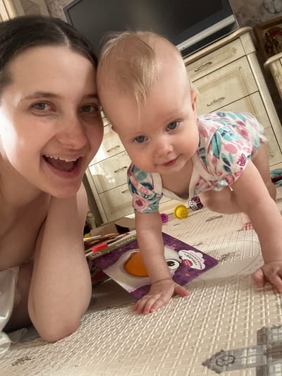 A woman and a baby are posing for a selfie. They are indoors, lying on a white textured playmat with grey outlines. The baby, wearing a floral patterned outfit, is positioned above the woman, their heads touching. The woman is smiling and looking into the camera. To the side, there is a colorful children's book with text in Russian. There is also a toy, and a white cabinet. It is not possible to determine the location in Pokrov, Russia from this image.