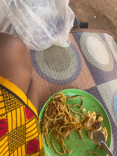An overhead view, captured in Oturkpo, Nigeria, depicts a meal in progress. In the lower-left quadrant, a person's bare forearm and thigh with brown skin are visible, partially covered by a yellow fabric featuring red and black geometric patterns. A round, bright green plastic plate occupies the center-right, holding partially consumed spaghetti mixed with a red-brown sauce and solid food fragments. A silver-colored metal fork is actively lifting a portion of the spaghetti from the plate. The plate and the person's leg rest on a woven mat displaying a repetitive pattern of intertwined circles and squares in shades of beige, brown, and purple. A clear, crumpled plastic bag containing indistinct items is positioned on the mat in the upper-center.