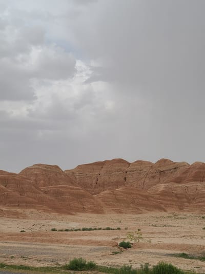 The image shows a geological formation in Kuchlak, Pakistan under a cloudy sky. The formation is characterized by reddish-brown, layered rock, possibly sedimentary in nature, with significant erosion creating ridges and gullies. Vegetation, including grass and small bushes, is sparse but present along the base of the formation. The sky is overcast, obscuring details of the horizon and suggesting potentially inclement weather. The photograph is taken from a ground-level perspective. No people are visible.