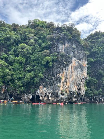 The image captures a vertical view of towering limestone cliffs covered in lush green foliage, characteristic of the karst landscape in Khlong Khian, Thailand. The cliffs rise dramatically from a calm, emerald-green body of water. Along the base of the cliffs, a line of colorful kayaks is tethered together, suggesting a tour or recreational activity. Several people are visible, some in the kayaks and others appearing to be standing on a narrow ledge at the waterline, possibly preparing to embark or disembark. The water reflects the sky and the surrounding cliffs, creating a shimmering effect. The sky above is partly cloudy, with streaks of white clouds against a blue expanse. The lighting suggests it is daytime, likely late morning or afternoon, with soft, diffused sunlight. The scene conveys a sense of natural beauty and adventure. There is no visible text in the image.