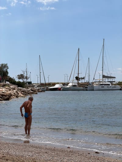 The image depicts a scene in Glifada, Greece, featuring a beach and marina. In the foreground, a man is standing in the shallow water near the shoreline. He is wearing blue swimwear. The beach is composed of small pebbles.
In the background, multiple sailboats are docked in a marina. The masts of the sailboats extend high into the clear, blue sky, which has a few scattered clouds. The shoreline includes trees, bushes, and some rocks and small stones.