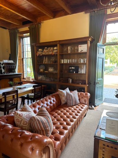 This is an interior shot. A large, tufted brown leather sofa sits next to a wooden display cabinet filled with glasses, dishes, and decorative items. A dining table with chairs is placed alongside the sofa. Light streams in through a door leading to the outside, and a window shows foliage beyond. The interior is decorated with wood panelling and green curtains. Given the architectural style and interior decor, it's likely that this photo was taken in a heritage building in Stellenbosch Nu, South Africa. The setting suggests a well-maintained establishment, possibly a historic hotel or restaurant.