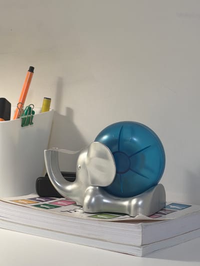 The image depicts a close-up view of a desk or shelf, with a white wall in the background. In the foreground, a stack of books forms the base. On top of the books rests a silver elephant-shaped tape dispenser, with its trunk holding a piece of tape. Behind the tape dispenser, a translucent blue spherical object, possibly a stress ball or a toy, is visible. To the left of the elephant is a white cylindrical container holding various writing utensils, including an orange marker, a yellow marker, and a black marker, along with a green binder clip. The scene appears to be set in an indoor environment, likely an office or study, in the city of Kfar Saba, Israel. There are no people or discernible actions taking place, and the focus is on the arrangement of desk objects.