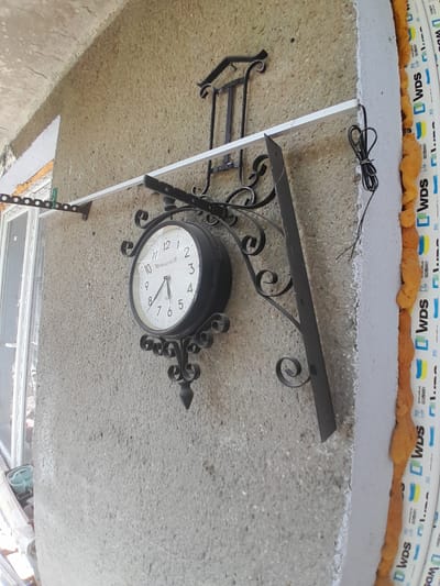 This media file shows a close-up, slightly angled view of a black, ornate, double-sided clock mounted on a concrete surface. The clock face is white with black numerals and hands, indicating approximately 10:10. The text "NEW YORK ELECTRIC CORP." is visible on the clock face, along with what appears to be a serial number or model number. Decorative black metal scrollwork extends from the mounting bracket to frame the clock. Above the clock, there is a white band with blue and yellow markings and the text "WDS" and "ISO 9001" repeated, suggesting it is related to window manufacturing or installation. A black electrical cord is coiled on the concrete surface above the clock. The concrete surface appears weathered. The scene suggests an outdoor setting, possibly a building facade or overhang. The lighting is consistent with daytime.