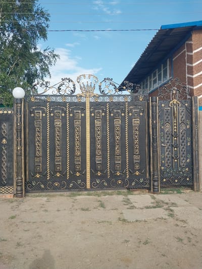 The image shows a section of a decorative metal gate in Aqsu city administration, Kazakhstan. The gate is primarily black with intricate gold accents. It features a repeating pattern of rectangular panels with various designs, including what appears to be a stylized X pattern, floral motifs, and decorative borders. The metalwork is ornate, with scrolling elements and detailed embellishments.
The gate is positioned outdoors, adjacent to a building with a brown brick facade and a blue overhang. To the left of the gate, a portion of the sky with white clouds is visible, along with the branches of green trees. A white spherical light fixture is attached to the gate. The ground in front of the gate is a light-colored, somewhat uneven surface, possibly dirt or gravel, with sparse patches of green grass. The orientation of the image suggests it might have been taken during the daytime, likely with clear or partly cloudy weather. There is no visible text in the image.