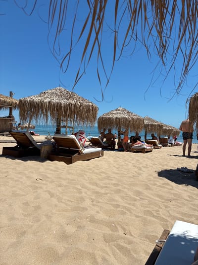 This is a photograph of a beach scene in Shkorpilovtsi, Bulgaria, on a bright, sunny day. The foreground is dominated by sand and several sun loungers. In the middle ground, there are multiple thatched umbrellas providing shade for more loungers. People are seen relaxing on the loungers, some partially obscured by the umbrellas. In the background, the sea meets the horizon under a clear blue sky. A beach bar structure is visible on the left side of the image.
