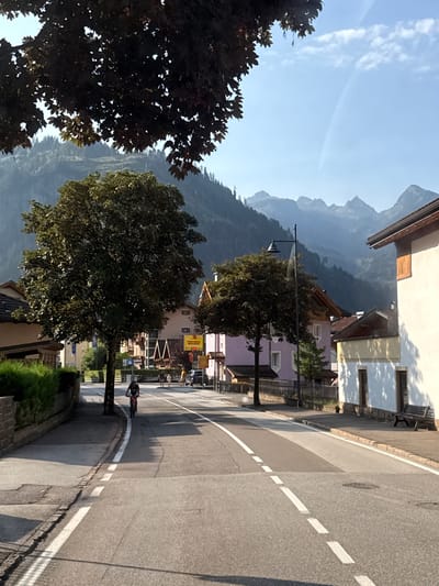A wide shot captures a street scene in the town of Predazzo, Italy. The foreground features an asphalt road with white lane markings. A lone cyclist, wearing a dark shirt and shorts, rides a bicycle towards the viewer. To the left of the road is a paved sidewalk bordered by a stone wall and a well-maintained hedge. Further left, a large tree with dark green foliage dominates the mid-ground.
On the right side of the street, several buildings are visible. The closest building is a white structure with a dark roof and a wooden accent above a window. A dark wooden bench sits on the sidewalk in front of this building. Behind it, other houses with pitched roofs and light-colored facades line the street.
In the background, more houses are visible, including a pink building with dark trim and a building with a yellow facade. A streetlamp stands near the pink building. Beyond the town, the scene transitions to a mountainous landscape. Lush green forests cover the lower slopes of the mountains, which rise into jagged, misty peaks against a clear blue sky. The overall atmosphere is serene and sunny, suggesting a pleasant day in a mountainous Italian town.