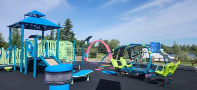 This is a medium-angle, wide shot of a modern, vibrant playground on a clear, sunny day. The playground features a multi-level structure with a prominent blue canopy adorned with white butterfly and floral patterns. The main structure is a combination of blue and bright green metal and plastic elements, including slides, climbing nets, and railings. There are also swings, a unique spinning apparatus with green seats, and a pink archway in the background. The ground surface is black rubberized material with colorful circular and oval-shaped inlays. Trees and other foliage surround the playground, and a body of water, likely a lake, is visible in the background with a wooded shoreline. The sky is a bright blue with scattered white clouds, suggesting it is daytime. The scene appears to be a public park in Edmonton, Canada. No people are prominently featured, though a few figures can be seen faintly in the background near the swings. The overall impression is a well-maintained and inviting outdoor recreational space.