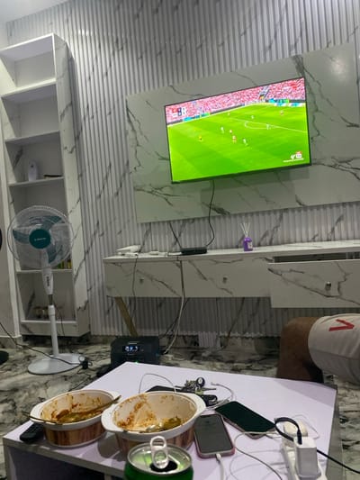 A room displays a TV showing a soccer game. The TV is mounted on a marble-patterned wall. Below the TV is a white marble shelf and a white bookshelf with a fan. A white table sits to the right, cluttered with phones, chargers, keys, dirty serving dishes, and a soda can. A person’s leg is visible next to the table.