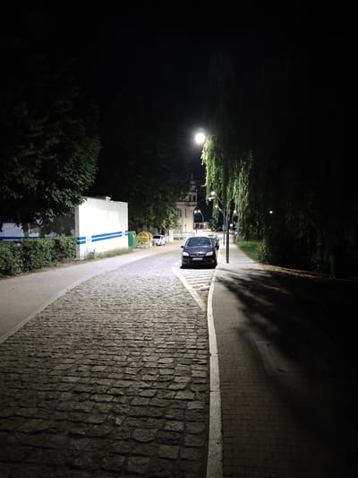 The image depicts a street scene at night in Stargard, Poland, likely during summer due to the lush foliage. A cobblestone path occupies the foreground, curving to the left and illuminated by artificial light. To the left of the path is a white building with blue horizontal stripes and a hedgerow. Further down the street, a dark-colored sedan is parked along the right side of the road, which appears to be paved with asphalt. A streetlamp casts light on the parked car and the adjacent sidewalk. Tall trees with dense foliage line the right side of the street, their branches casting shadows across the sidewalk. In the background, a building with a steeple is visible, suggesting a historical or civic area. The overall atmosphere is quiet and somewhat isolated due to the darkness and sparse human presence.