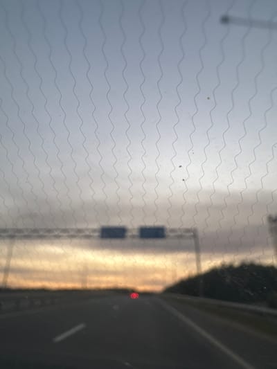 The image is a photograph taken from inside a vehicle, looking out through a patterned windshield. The scene depicts a highway at sunset. The sky is a gradient of pale blue and gray at the top, transitioning to warm orange and yellow hues near the horizon, suggesting either sunrise or sunset. A line of dark trees is visible on the right side of the road, partially obscuring the horizon. In the distance, an overhead highway sign with two rectangular blue panels is blurred by the windshield's pattern and the image's focus. The highway itself is dark asphalt with a white lane marker visible on the left side. A single red light, possibly from another vehicle, is visible in the middle distance on the right side of the road. The windshield has a repeating wavy line pattern, which is superimposed over the entire scene. The image is taken in or near Mstikhino, Russia, as indicated by the prompt.