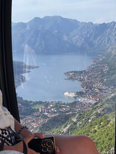 The image displays a high-angle panoramic view of a large bay and coastal town, observed from an elevated position through a window. The window's dark frame is visible on the left and bottom, exhibiting reflections including vertical light streaks and partial images of the foreground subject.

The central feature is a wide, calm body of dark blue water, identified as a bay or fjord, surrounded by steep, rugged mountains. The mountains exhibit green foliage on their lower and mid-slopes, transitioning to grey rocky terrain at higher elevations, and appear lighter blue due to atmospheric haze in the distant background.

Along the bay's edge, a densely populated urban area is visible, characterized by numerous buildings, many with red-tiled roofs, sprawling along the coastline and ascending the lower mountain slopes. A large, white multi-deck cruise ship is prominently docked at a pier in the bay's foreground-right section. Adjacent to the cruise ship is a marina filled with various smaller boats and yachts. Several motorboats are seen navigating the bay, leaving distinct white wakes.

In the lower-left foreground, the partial figure of a person is visible, including their forearms and upper thighs. The person wears a white garment and a black-and-white patterned fabric. A blue beaded bracelet is present on their left wrist. Their right hand holds a gold-toned smartphone, the rear of which displays a prominent multi-lens camera array.

This scene is located in the city of Mirac, Montenegro.