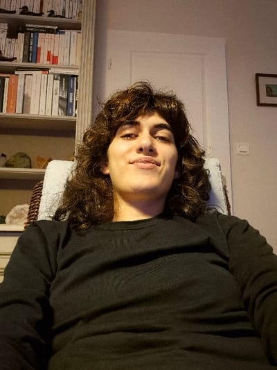 The image depicts an individual seated indoors in Châtelaillon-Plage, France. A young adult, with shoulder-length dark curly hair and fair skin, is centrally positioned, facing the camera with a slight smile. The individual wears a black, long-sleeved crew-neck shirt and is seated in a chair with a visible light-colored cushion and a brown wicker-like frame.
To the left of the individual, a light-colored wooden bookshelf occupies the background. The bookshelf contains multiple rows of books of varying sizes and colors, along with several decorative items including small dark figurines (possibly ducks) on the top shelf and what appear to be geological specimens (rocks/crystals) on lower shelves.
To the right of the individual, a light-colored wall is visible. A white closed door with a doorknob is present on the right side of the frame. Below and to the right of the door, a white rectangular light switch is mounted on the wall. Further right and above the switch, a small, dark-framed picture with a light-toned image hangs on the wall. The scene is illuminated by an unseen light source, casting soft shadows.