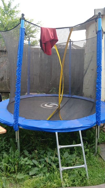 In Kolyvan, Russia, a trampoline with a black safety net and blue padding is situated outdoors. A red garment is draped over the top of the net, and a yellow strap hangs down inside. A silver ladder leans against the side of the trampoline, which is surrounded by green foliage and grass. A wooden fence is visible in the background, along with a section of a building. The sky above is overcast and white.