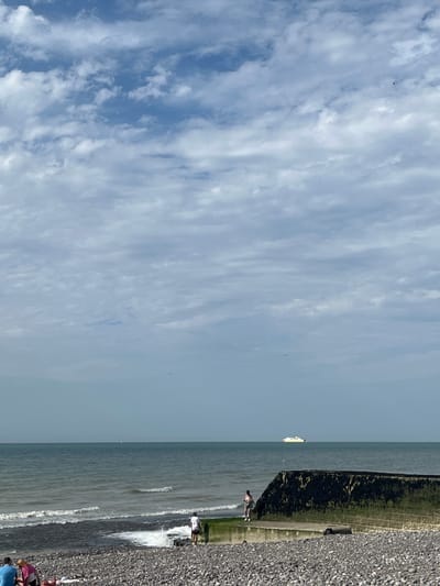 This is a vertically oriented, eye-level, medium shot of a cloudy sky and a beach scene. The sky fills the majority of the left side of the frame, with wispy, white clouds streaking across a pale blue canvas. A tiny speck in the sky might be a bird or a distant aircraft. On the right side of the image, a pebble beach is visible, meeting the calm grey ocean. The water has gentle waves breaking on the shore.

Along the water's edge, a few people are present. One person is wading in the shallow water, and a group of people, some wearing colorful clothing, are gathered on the beach. A white ferry or ship is visible in the distance on the horizon, on the left side of the frame. A dark, textured structure, possibly a sea wall or breakwater, runs along the edge of the beach. The light suggests it is daytime, likely mid-morning or afternoon, with overcast conditions. The overall mood is serene and casual, indicative of a beach outing. No text is visible in the media file.