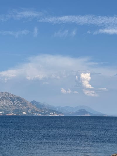 An expansive view of the Adriatic Sea stretches across the foreground, its deep blue waters rippling gently under a bright sky. In the mid-ground, a rugged coastline is visible, characterized by steep, mountainous terrain dotted with scattered white buildings, indicative of a town or village. The mountains appear to be covered in sparse vegetation, with rocky outcrops exposed on their slopes. Further in the distance, a series of hazy, blue-hued mountains recede into the horizon. The sky above is a clear blue, punctuated by a scattering of white clouds. To the right, a large, puffy cumulus cloud stands out, with thinner, wispy clouds streaking across the upper portion of the sky. The scene captures a coastal landscape under a predominantly clear sky with some cloud formations. The location appears to be near Dubraljevina, Montenegro.