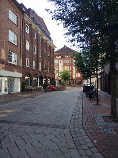 The image shows a street scene in Cuxhaven, Germany, likely in the historic center. On the left, a multi-story brick building with white-framed windows lines the street. Below, a ground-floor establishment with white doors and windows is visible. Across the street, another building features a prominent "HOTEL" sign and a similar architectural style. A car is parked on the right side of the street, next to several bicycles locked to a lamppost. The street is paved with cobblestones and bricks, and trees are planted along the sidewalk. Outdoor seating for a cafe is set up in front of the brick building on the left. The lighting suggests it is daytime, with the sun casting shadows.