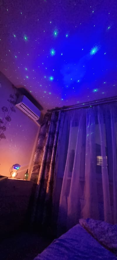 The image depicts an indoor scene with a ceiling illuminated by a star projector, creating a starry night sky effect with blue and green lights. A white air conditioner unit is mounted high on a wall to the left. To the right, sheer purple curtains are drawn over a window. A patterned curtain is partially visible to the left of the sheer curtains. On a surface in the lower left, a lamp with a blue light is visible, along with a green bottle. The overall lighting is dim and atmospheric, with a focus on the projected stars on the ceiling. The location is identified as Romashkovo, Russia.