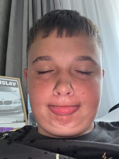 A close-up selfie shows a young boy with his eyes closed and his tongue sticking out. He is wearing a black t-shirt. In the background, a framed picture of a car is visible, with the name "SLAV" printed on it. The environment suggests an indoor setting with curtains in the background.
