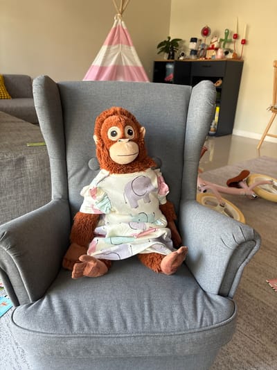 A plush orangutan toy is seated upright in a light gray upholstered armchair, centrally positioned within what appears to be a domestic interior located in Bang Kaeo Subdistrict, Thailand. The orangutan, featuring brown fur, large brown eyes, and a light-colored muzzle, wears a loose-fitting, short-sleeved garment patterned with light pastel elephants and abstract shapes. Its feet are positioned on the chair's seat cushion.
To the left and slightly behind the armchair, a section of a gray fabric sofa with a yellow decorative pillow is visible. In the background, against a light-colored wall, a pink and white striped play teepee stands. Further to the right in the background, a dark gray cabinet with a wooden top displays various small objects, including a plant, a baby monitor, and decorative figurines. A wooden balance bike with yellow wheels and a brown seat lies on its side on a light gray patterned rug on the floor to the right of the armchair. The room exhibits natural ambient lighting.