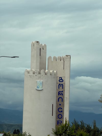 The image depicts a white, castle-like tower against a cloudy sky, likely located in or near Mrgvali Chala, Georgia, given the Georgian text "მესტია • სოფელი" ("Mestia • Village") displayed in blue letters on the tower's facade. A coat of arms is also visible on the tower. Green foliage is present to the right. A streetlight pole is visible at the bottom left.