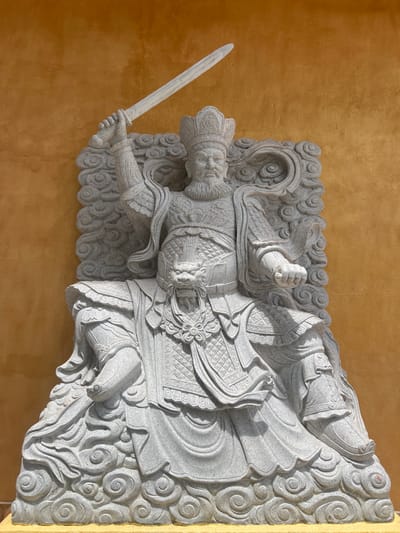 A grey stone statue of a male figure is depicted against a textured, plain yellow-orange wall in Đăk Mar, Vietnam. The figure, possibly a deity or warrior, wears an ornate crown and elaborate, patterned armor, including a prominent dragon head embellishment on the chest. It possesses a full beard, mustache, and a stern facial expression. The statue is rendered in a seated or poised posture, with its right arm raised high, gripping a carved stone sword pointing upwards and to the left. The left hand is clenched into a fist. The figure is flanked by and rests upon intricately carved cloud-like patterns, which form part of its base and surround its form. Shadows suggest outdoor lighting conditions.