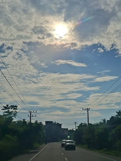 The image depicts a view from a vehicle on a paved road in Keude Lueng Putu, Indonesia, with lush green trees lining both sides of the road. Utility poles and power lines are prominently visible overhead, stretching across the frame. The sky is a vibrant blue, adorned with a mix of fluffy white clouds and some darker, scattered clouds. The sun is high in the sky, partially obscured by clouds, creating a bright glare and a faint rainbow effect. In the distance, a dark, multi-story building is visible. Two cars are traveling on the road in the same direction as the viewer. The scene suggests a daytime journey on a road surrounded by natural foliage and infrastructure.