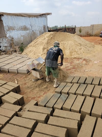 The image depicts an outdoor construction site in Nwofia, Nigeria. A male individual, viewed from behind, is centered in the midground, engaged in manual labor. He wears a dark t-shirt with a light graphic (text partially visible as "BADGER WINGS"), green shorts, and light-colored shoes with socks, topped with a light-colored cap. He is bent forward, holding a silver-bladed shovel, actively working with sand or other material on the dirt ground.
In the immediate foreground and extending to the left, numerous rectangular concrete blocks are systematically arranged on the bare earth, some stacked, others laid flat. To the left of the individual, a metal brick mold and a white plastic bag are visible on the ground. Directly behind the worker is a large, light-colored mound of sand.
In the background, a dilapidated structure made of corrugated metal sheets stands on the far left. Further right, behind the sand pile, a wooden gate or truck bed structure is partially visible. A concrete block wall extends from the right background towards the center, with a small plant growing nearby. A portion of a light-colored vehicle is visible on the far right. The sky overhead is light and overcast. The ground throughout the scene is composed of dry dirt and sand.