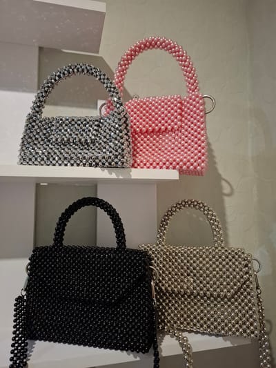 Four beaded handbags are displayed on white shelves against a textured, light-colored wall. The top shelf holds a silver, faceted beaded handbag to the left and a pink, round beaded handbag to the right. The bottom shelf displays a black, round beaded handbag on the left and a silver, round beaded handbag on the right. The handbags have rounded handles. The scene appears to be an indoor retail display, possibly in a store located in Korenovsk, Russia. No people are visible.
