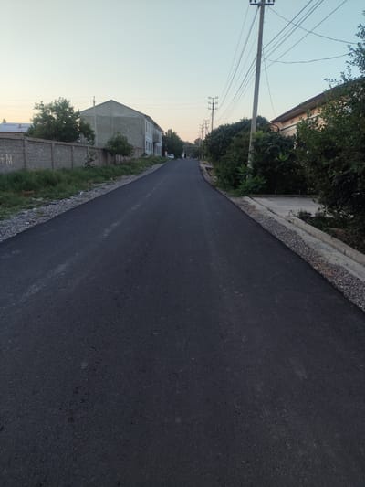 The image shows a paved road in the city of Dangir, Uzbekistan. The road is dark asphalt and appears to be recently paved. On the left side of the road, there is a low brick wall with overgrown grass and small trees. Beyond the wall, there is a building with a light-colored facade. On the right side of the road, there are trees and a concrete curb. In the background, there are power lines and utility poles. The sky is a pale blue with some haze. There are no people visible in the image. The overall impression is one of a quiet, somewhat rural street.