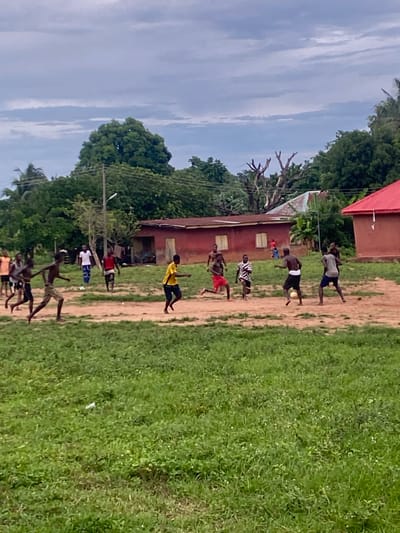 The image captures an outdoor scene in Iyare, Nigeria, under a uniform overcast sky with visible stratiform clouds. A group of approximately fifteen young males is present on a playing area composed of reddish-brown dirt paths and patches of green grass. The primary activity is a football (soccer) game, with a light-colored football situated centrally on the field. One male, seen from behind wearing dark shorts with a white waistband and a dark tank top, is in the process of kicking the ball. Another male in a grey t-shirt and dark shorts is positioned opposite him, facing the kick. Other participants, dressed in various casual attire including red shorts and a red tank top, a striped shirt, a yellow t-shirt, and white shirts with patterned trousers, are distributed across the field, either running, observing, or moving towards the play.

In the background, a cluster of single-story buildings with red and metallic corrugated roofs is visible, partially obscured by dense green tropical foliage and trees. Notably, a prominent red-roofed building stands to the right, while another red-walled structure is positioned further left. A wooden utility pole with overhead wires extends across the mid-ground, accompanied by a street light pole. The foreground consists of more expansive green grass, within which a small, white piece of debris is visible in the lower-left quadrant.
