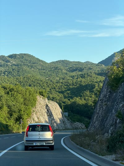 An eye-level, medium shot captures a silver Fiat Punto driving on a winding road with lush green mountains in the background. The car, seen from behind, has its brake lights illuminated. The road is asphalt with white lane markings. To the right of the road, a steep, rocky embankment is visible, covered with sparse vegetation. To the left, the terrain slopes upwards, also covered in dense greenery. The sky is clear and blue with a few wispy clouds. The scene appears to be in a mountainous area, possibly Vrela, Montenegro, given the context.