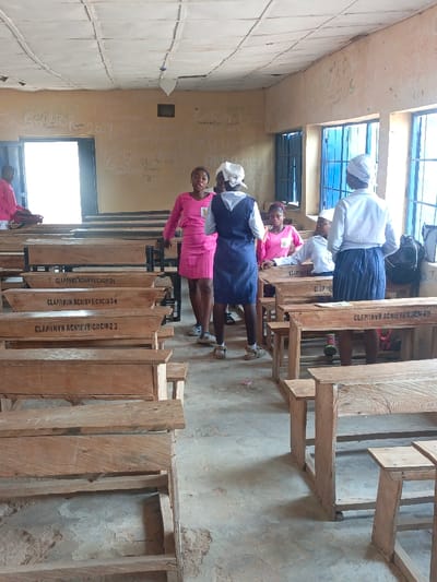 The image displays an indoor classroom located in Mada, Nigeria. Seven individuals, identified as female students, are present. The room features light-colored walls extensively covered with handwritten graffiti and inscriptions, a paneled ceiling showing signs of deterioration and discoloration, and an uneven concrete floor with visible wear. Multiple rectangular windows fitted with blue metal grates are situated on the right wall. A brightly lit open doorway is visible on the left wall.

Numerous wooden school desks and separate benches are arranged in rows. Several desks are stenciled with "CLAPHRVN ACHIEVE CICIO" followed by identification numbers (e.g., 21, 38, 23, 09). A black backpack rests near the windows on the right. A small red object is on the floor in the central foreground.

On the left, one student in a pink uniform is seated at a desk, facing towards the doorway. In the central area, a student in a bright pink dress stands, facing right with her mouth open, appearing to speak or sing. She is facing another standing student who wears a blue sleeveless tunic over a white shirt and a white headwrap. To their immediate right, two students are seated at a desk; one wears a pink uniform with a badge, and the other wears a white shirt. On the far right, a student with her back to the camera wears a white shirt, a dark pleated skirt, and a white headwrap, appearing to observe or address the seated students. The scene depicts an active classroom or group interaction.