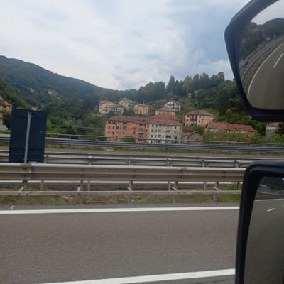 The image captures a view from a vehicle traveling on a highway, likely in Masone, Italy, as indicated by the presence of a road sign with Italian lettering. The scene is dominated by a highway with multiple lanes, separated by guardrails. In the background, a hillside is covered with dense green trees. Several buildings are visible on the slope, including apartment buildings and smaller houses, suggesting a residential area nestled into the landscape. A dark blue, rectangular signpost stands on the left side of the road, partially obscuring the view of the buildings on the hill. The sky is overcast with clouds. A side-view mirror of the vehicle is visible on the right, reflecting the road and the surrounding environment.