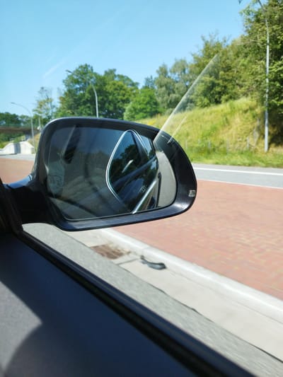 This media file captures a close-up view from inside a car, looking outwards through the passenger side window and the side mirror. The scene appears to be a daytime journey, likely a car ride, given the perspective and the reflection in the mirror.

The main subjects are the car's side mirror and a portion of the car's interior. The side mirror itself is a dark, possibly black, housing, and the mirror surface reflects the surrounding environment. Visible in the reflection is the interior of the car, including what appears to be a dashboard or seat. Outside the car, visible through the window and in the mirror's reflection, is a road with a distinctive red brick or paver surface lining the edge, adjacent to a lighter gray surface. Beyond the road, there is a green, grassy embankment covered in trees. The sky above is a clear, bright blue, suggesting fair weather and a sunny day. A light-colored line, possibly a road marking or a barrier, runs along the edge of the green embankment. A street light is also visible against the trees.

The activity taking place is a car journey. The perspective implies movement, as suggested by the slight blur in the background elements. The time of day is clearly daytime, with bright sunlight casting shadows and illuminating the scene. The weather appears to be pleasant and sunny. Location cues, such as the brickwork and the type of greenery, along with the provided location context of Bremerhaven, Germany, help to establish a specific setting. There is no visible text in the image. The overall impression is one of a routine car journey on a bright, clear day, with the focus on the car's external features and the passing scenery.