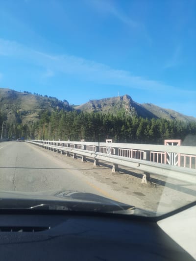 The image presents a scenic view captured from inside a vehicle, looking out onto a road winding through a mountainous and forested landscape. The upper portion of the frame is dominated by a clear, bright blue sky with a few faint, wispy clouds, indicating a sunny day.
To the left, rugged mountains rise, densely covered with dark green coniferous trees, likely pines. The forest extends up the slopes, and on a distant peak, a slender, light-colored structure, possibly a communication tower or monument, is visible against the sky.
A paved road, appearing to be a highway or main route, stretches into the distance, bordered on its left by a long, gray metal guardrail. This guardrail is supported by numerous posts, some of which feature light-colored or pale pink sections, adding a subtle visual detail. The ground immediately to the left of the road and beneath the guardrail is a dry, light brown dirt or gravel.
The right side of the image features a dark, curved section of a car's interior, likely part of the dashboard or door frame, confirming the perspective is from within a moving vehicle. The bright sunlight casts distinct shadows, highlighting the clear weather conditions. The overall scene suggests a journey through a picturesque natural environment, consistent with the mountainous and forested areas found in Katun', Russia. No discernible text is visible.
